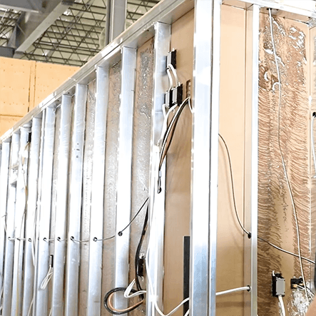 Interior view of an unfinished building wall with exposed metal studs and wiring. The structure is under construction, with visible insulation and wiring, in a well-lit industrial setting.