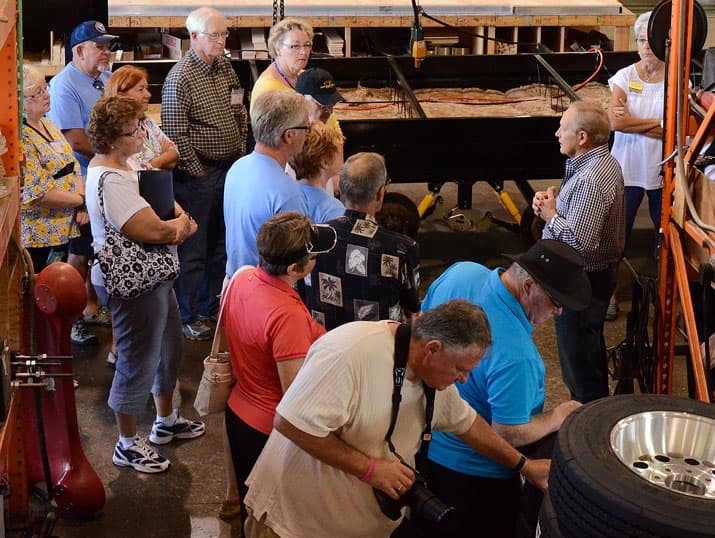 A group of people gathers in a workshop-like setting, listening to a man speaking. Various tools and equipment are visible, including a tire and machinery. Some individuals are taking notes or photos.