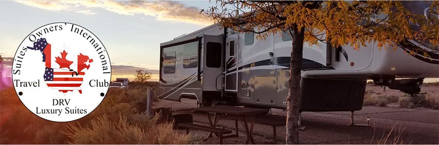 A luxury RV parked in a scenic area at sunset, with a picnic table beside it. On the left, a logo for the Suites Owners International Travel Club featuring an American flag shield and a maple leaf is visible.