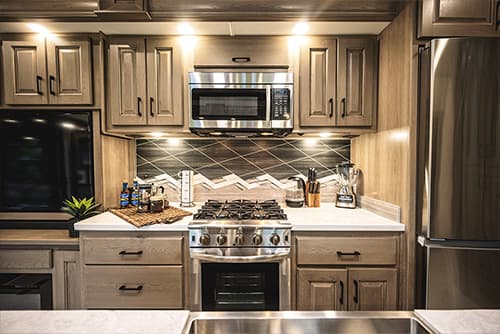 Modern kitchen with wooden cabinets, stainless steel appliances, and tiled backsplash. Features a stove with oven, microwave, and refrigerator. Countertop holds a blender, spice rack, potted plant, and a tray with drinks. Bright overhead lighting.